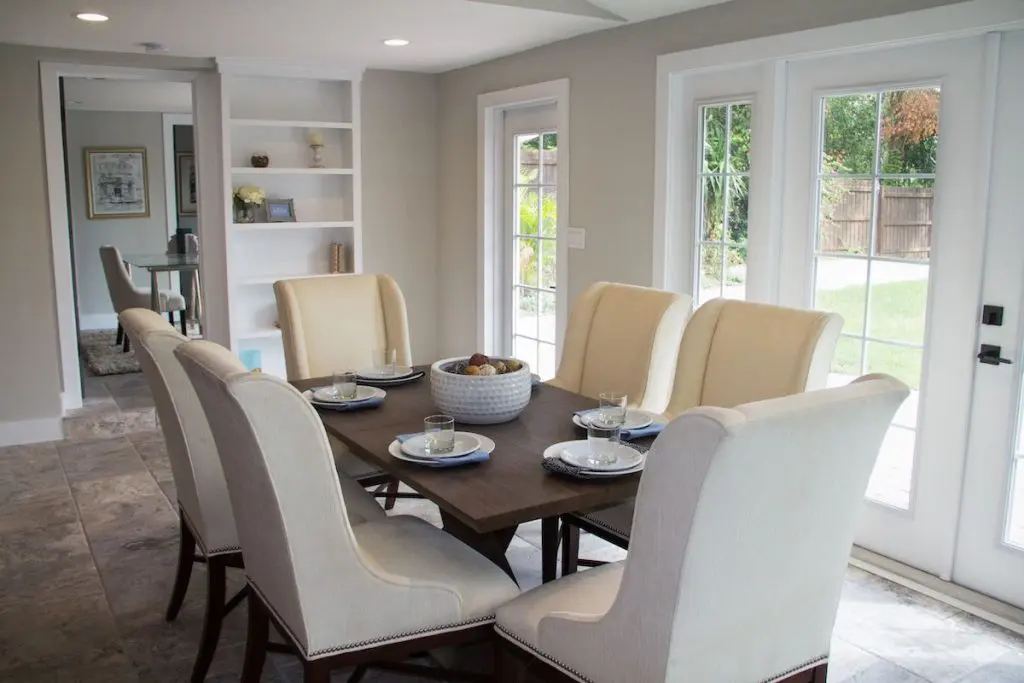Formal dining room of the house featured on Zombie House Flipping season two episode thirteen, containing French doors, a dining set with large neutral dining chairs, place settings, and a built in bookshelf nearby.