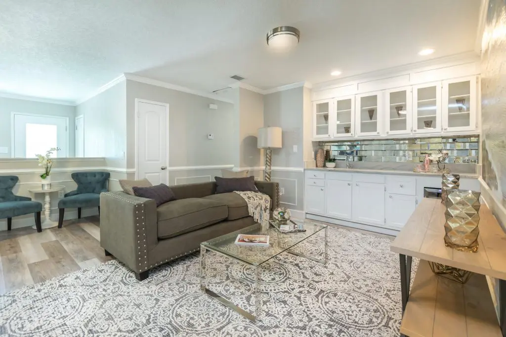 Living room with sofa, transparent coffee table, gray patterned rug, shelf display, and wetbar staged by MHM Professional Staging for Zombie House Flipping season three episode one.